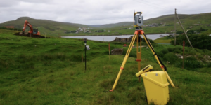 CSM SVQ EMN Plant Shetland Image of a theodolite planted in a field, pictured to the fore of the image, with earth moving equipment towards he rear left of hte image and with a small loch pictured in the centre of the image