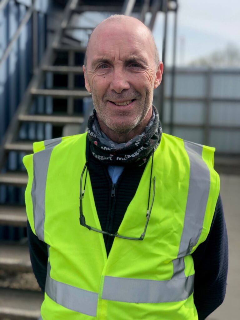 James Thomson featured in a yellow high vis  vest in profile smiling at the camera. Wearing a McTaggart Construction scarf round his neck