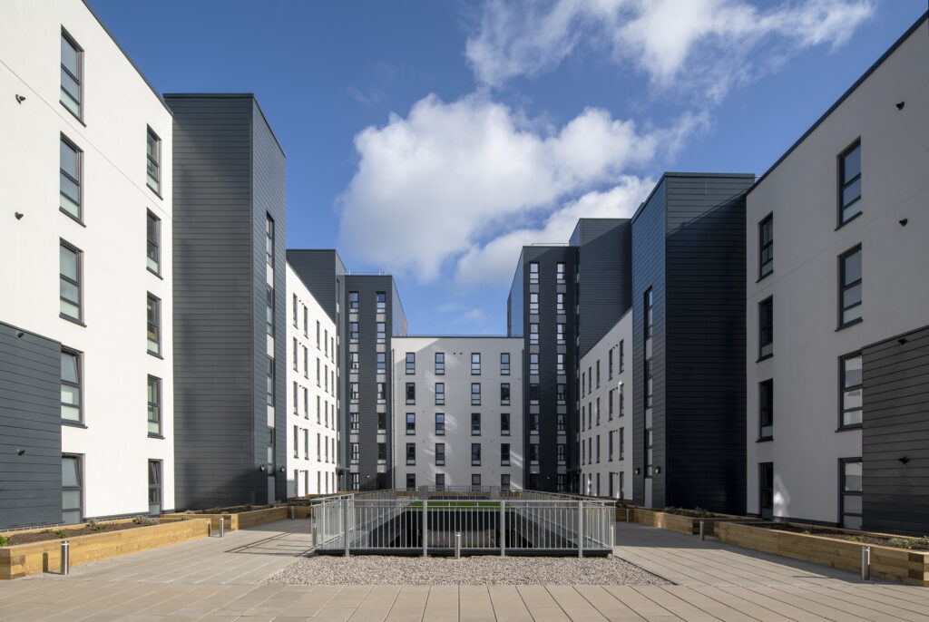 Surrounded by a modern designed white and grey block of flats. There is a courtyard in the middle with railings. There are wooden planterson the perimiter of the courtyard